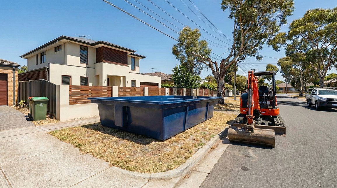 A dark blue fibreglass pool shell sitting on the front verge of a suburban Australian street next to a mini excavator, with a modern home and eucalyptus trees in the background 